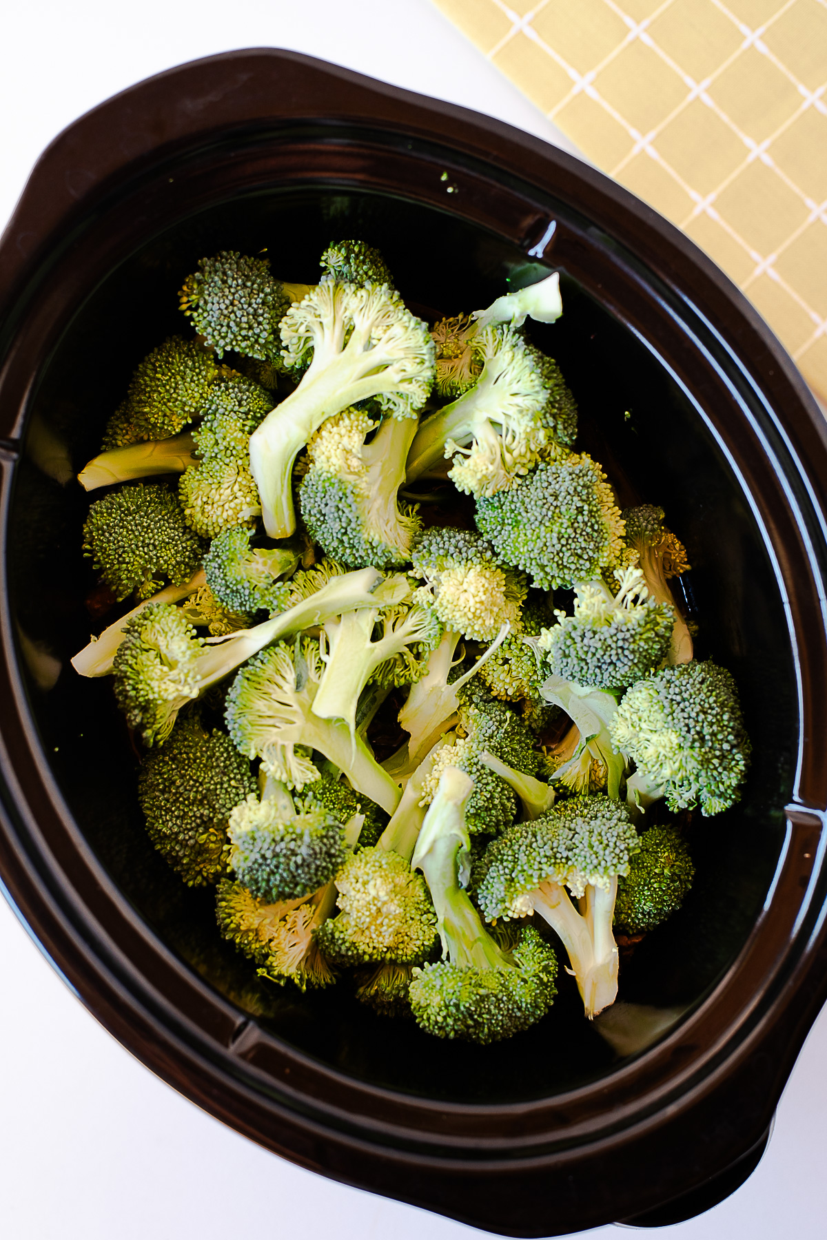 Overhead shot of raw broccoli florets layered on top of the cooked beef in the crockpot.
