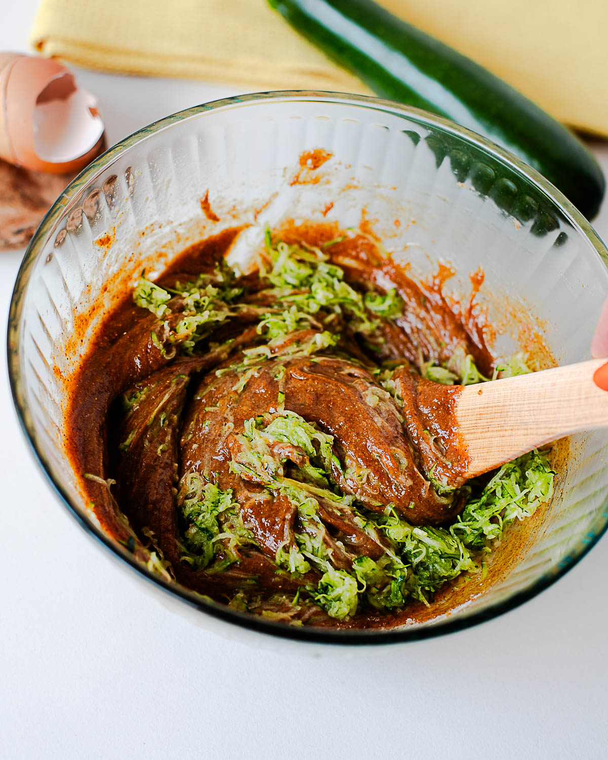 Shredded zucchini being folded into almond butter muffin batter with a wooden spoon