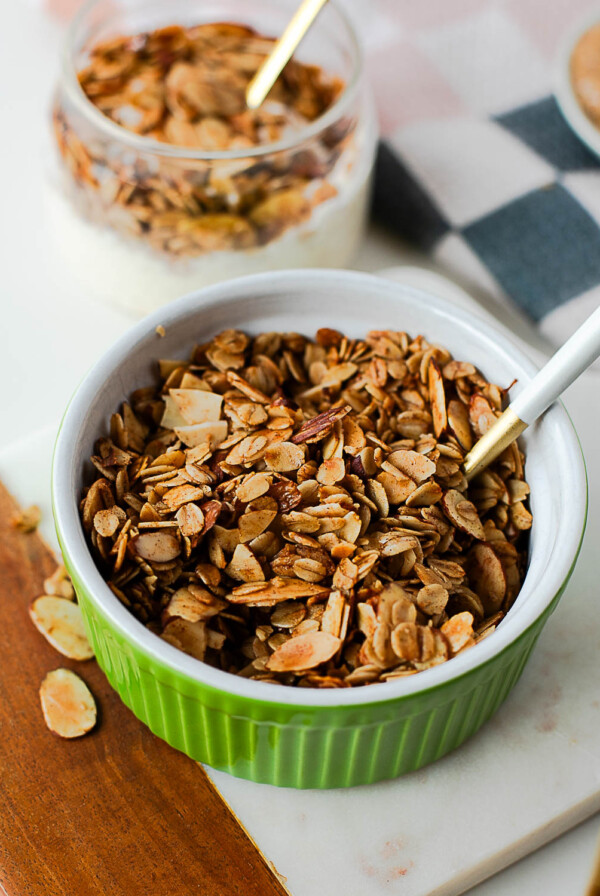 Green ramekin filled with golden toasted olive oil granola and a white spoon, surrounded by scattered oat and almond pieces.
