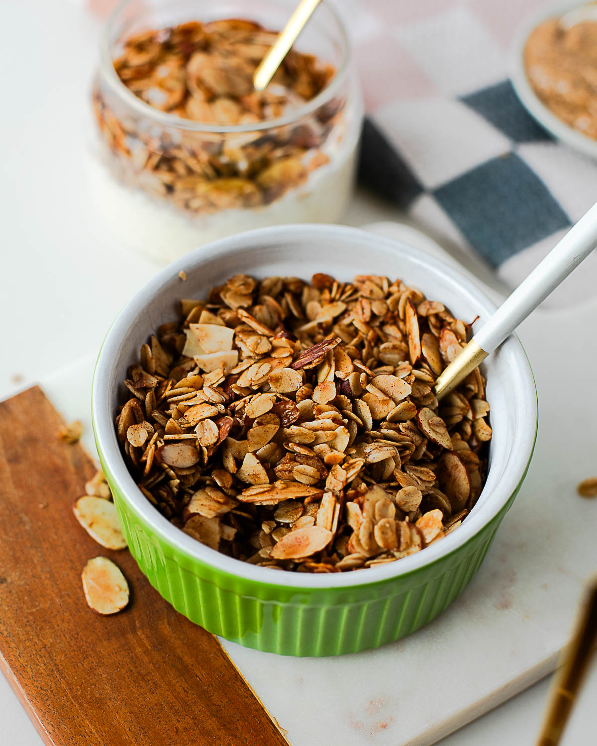 Green ramekin filled with golden toasted olive oil granola and a white spoon, surrounded by scattered oat and almond pieces.