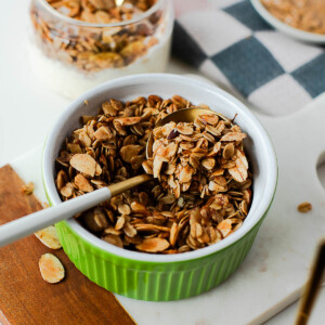 Close-up of almond maple olive oil granola in a green ramekin with a white spoon, showing the golden, toasty oats and almonds ready to serve.