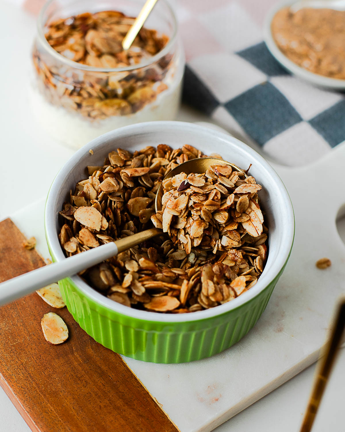 Close-up of almond maple olive oil granola in a green ramekin with a white spoon, showing the golden, toasty oats and almonds ready to serve.