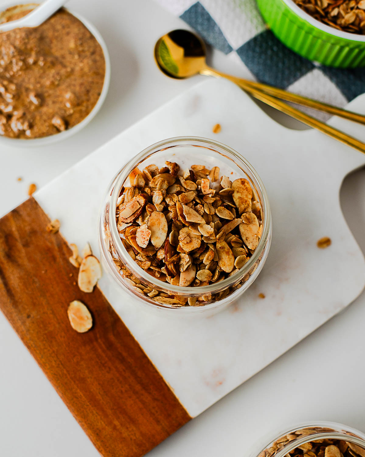 Overhead view of a glass jar filled with homemade olive oil granola on a marble and wood board, surrounded by almond butter and gold spoons for a cozy breakfast setup.
