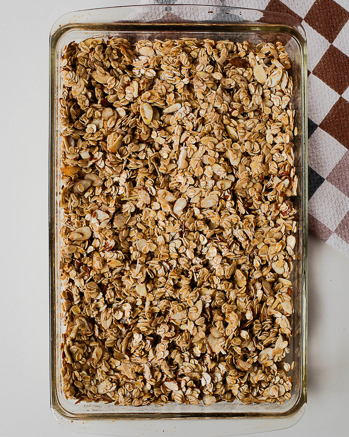 Overhead view of unbaked almond maple olive oil granola spread evenly in a glass baking dish, ready to go into the oven.