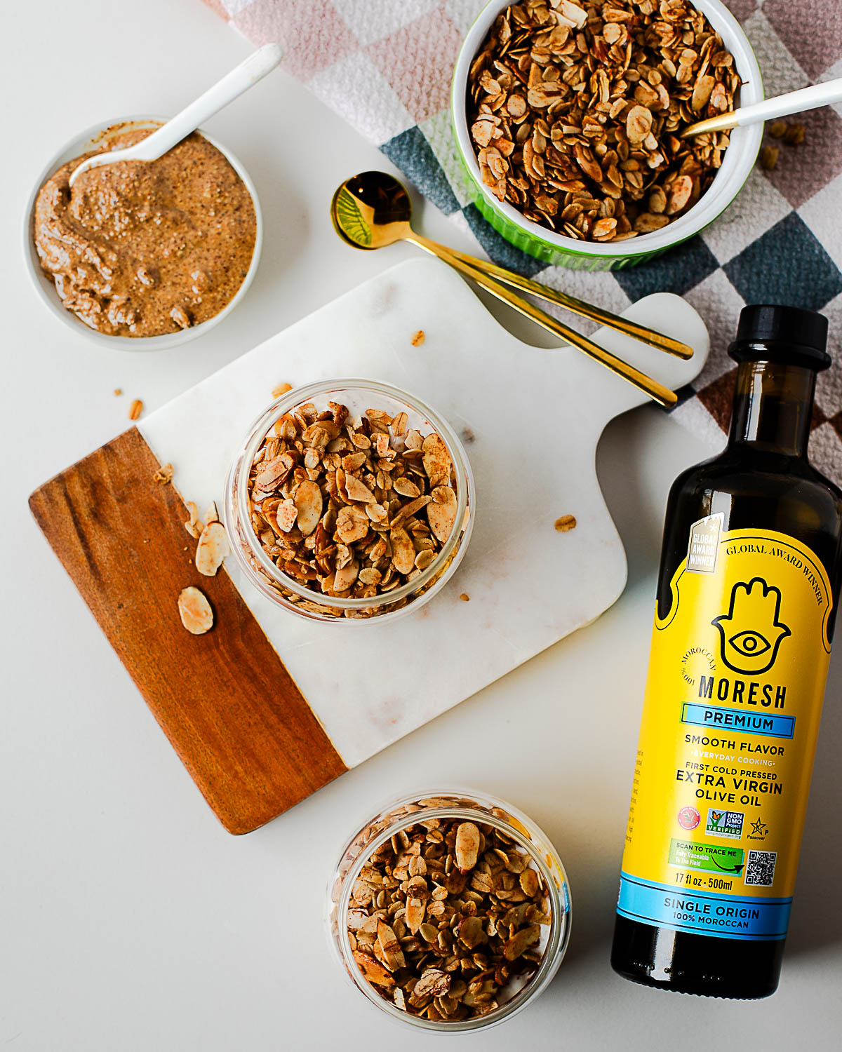 Overhead spread of breakfast setup with jars of granola, Moresh olive oil bottle, and gold spoons on a checkered napkin — cozy and inviting scene.
