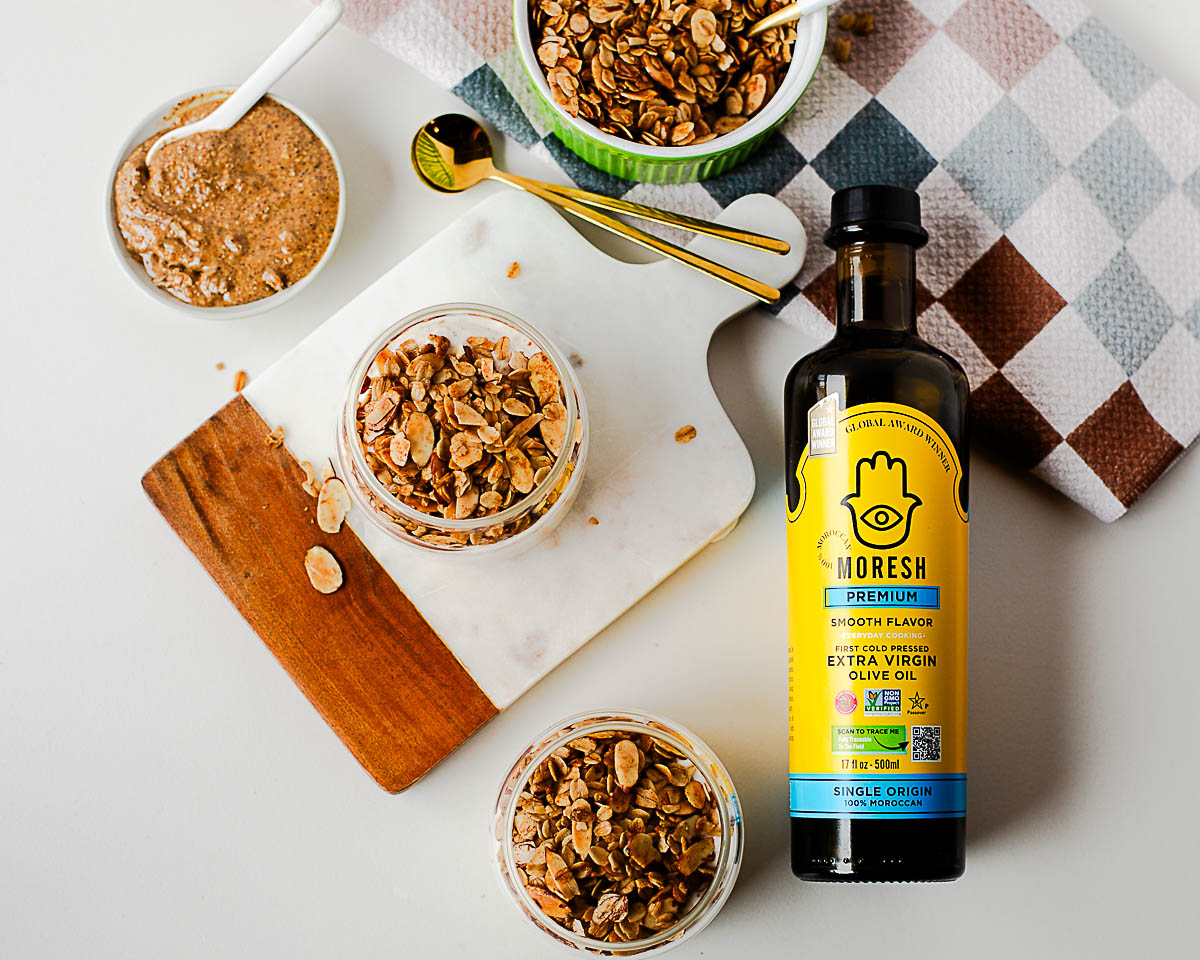 Overhead spread of breakfast setup with jars of granola, Moresh olive oil bottle, and gold spoons on a checkered napkin — cozy and inviting scene.