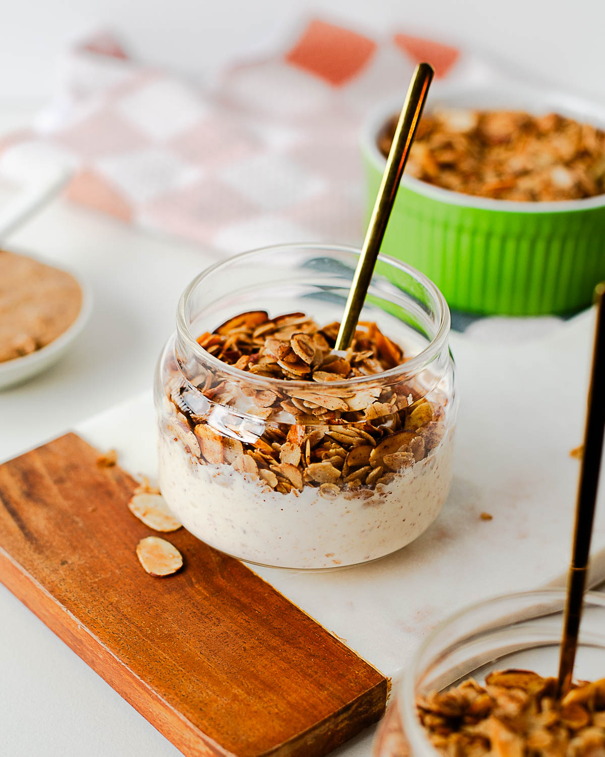 Side view of a glass jar filled with yogurt and homemade granola, with extra granola and almond butter in the background.