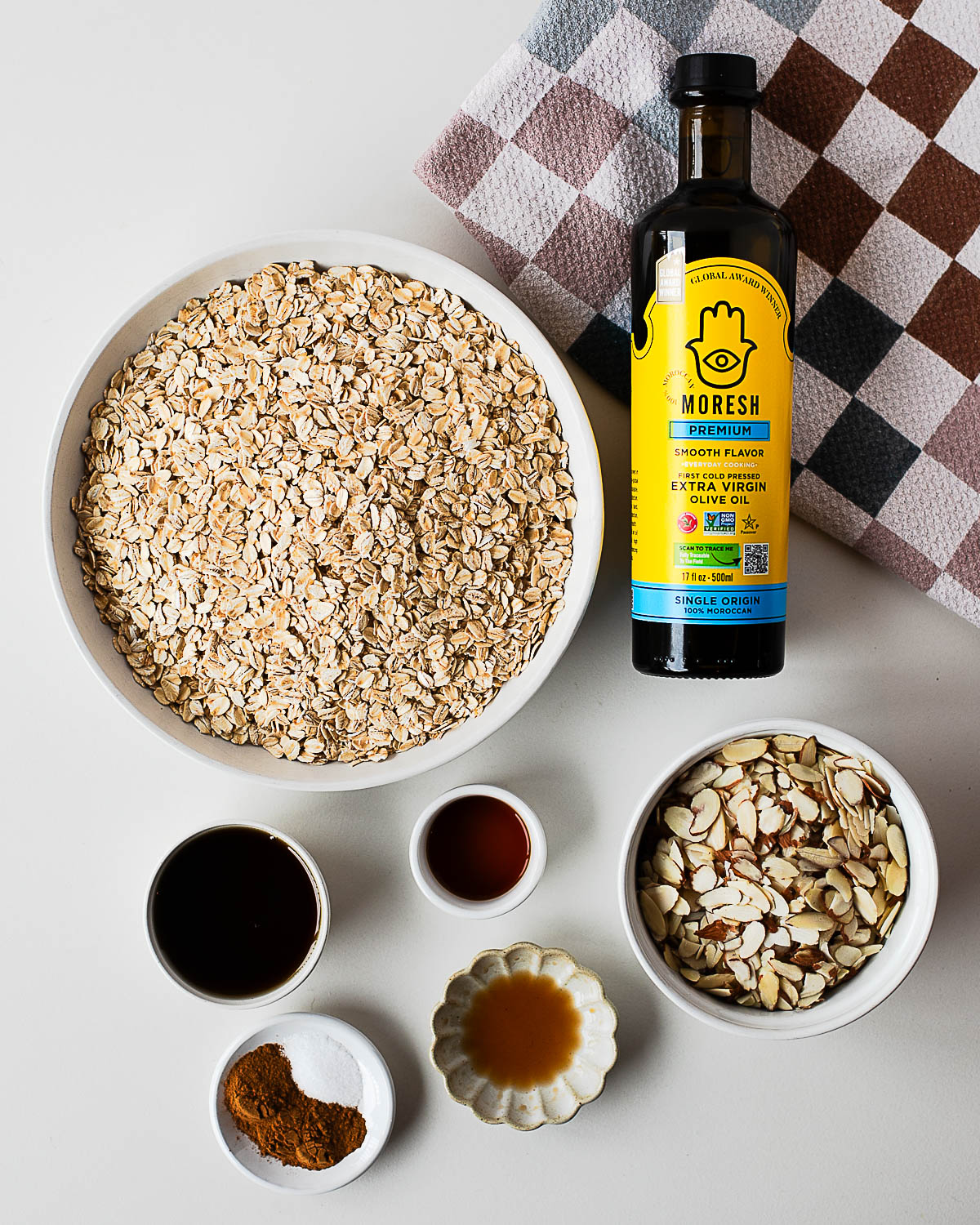 Overhead shot of the simple ingredients for almond maple olive oil granola: oats, sliced almonds, maple syrup, olive oil, cinnamon, and salt — with a bottle of Moresh olive oil featured beside them.