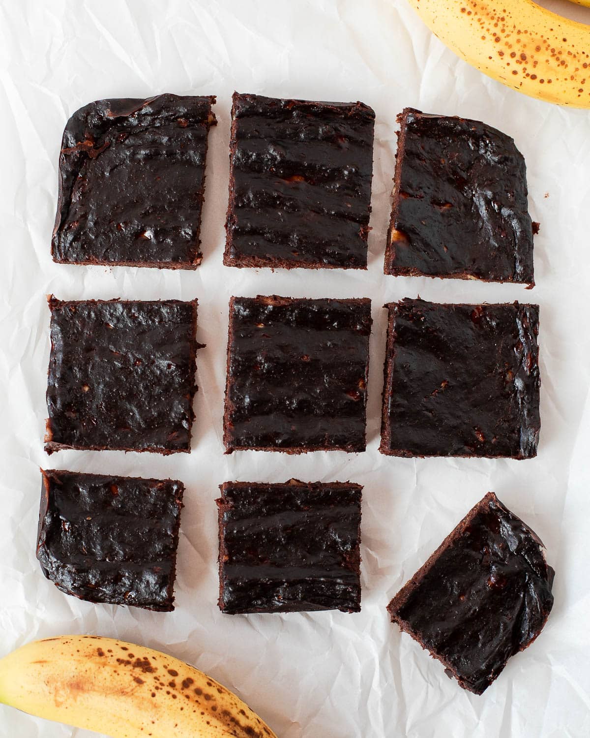 Overhead view of sliced 3 ingredient banana brownies arranged on parchment paper with ripe bananas.