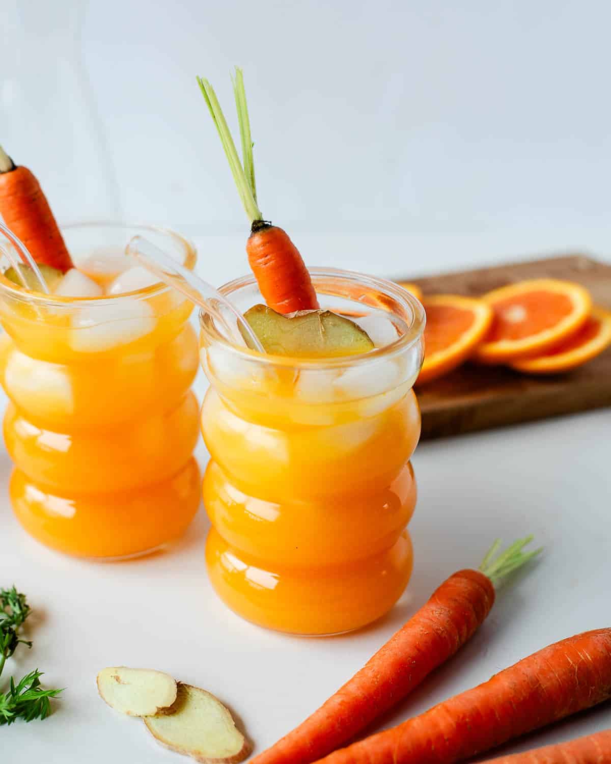 Two glasses of fresh carrot ginger juice with ice cubes, carrots, ginger slices, and oranges.