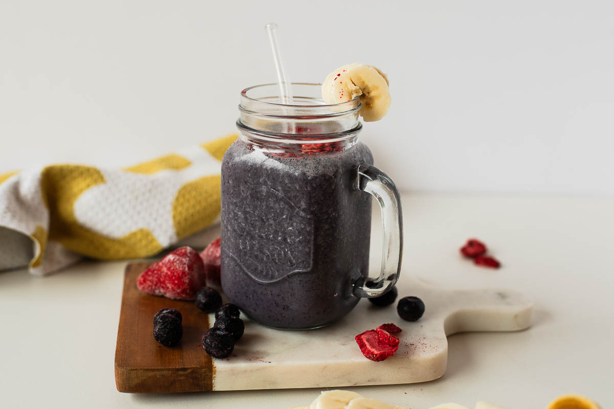 Mason jar of dark purple egg white protein shake topped with freeze-dried strawberries and a banana slice, set on a board with frozen berries.