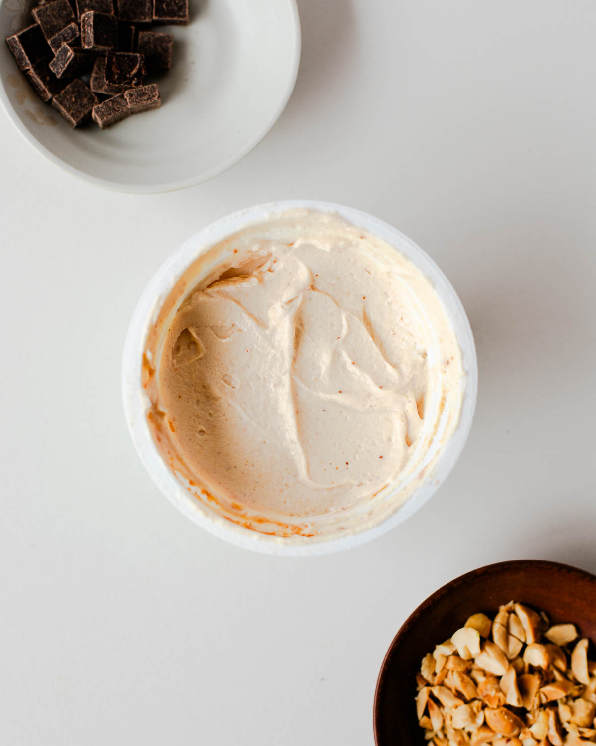 Greek yogurt stirred with peanut butter in a cup; dark chocolate chunks and chopped peanuts nearby for Snickers yogurt cup.