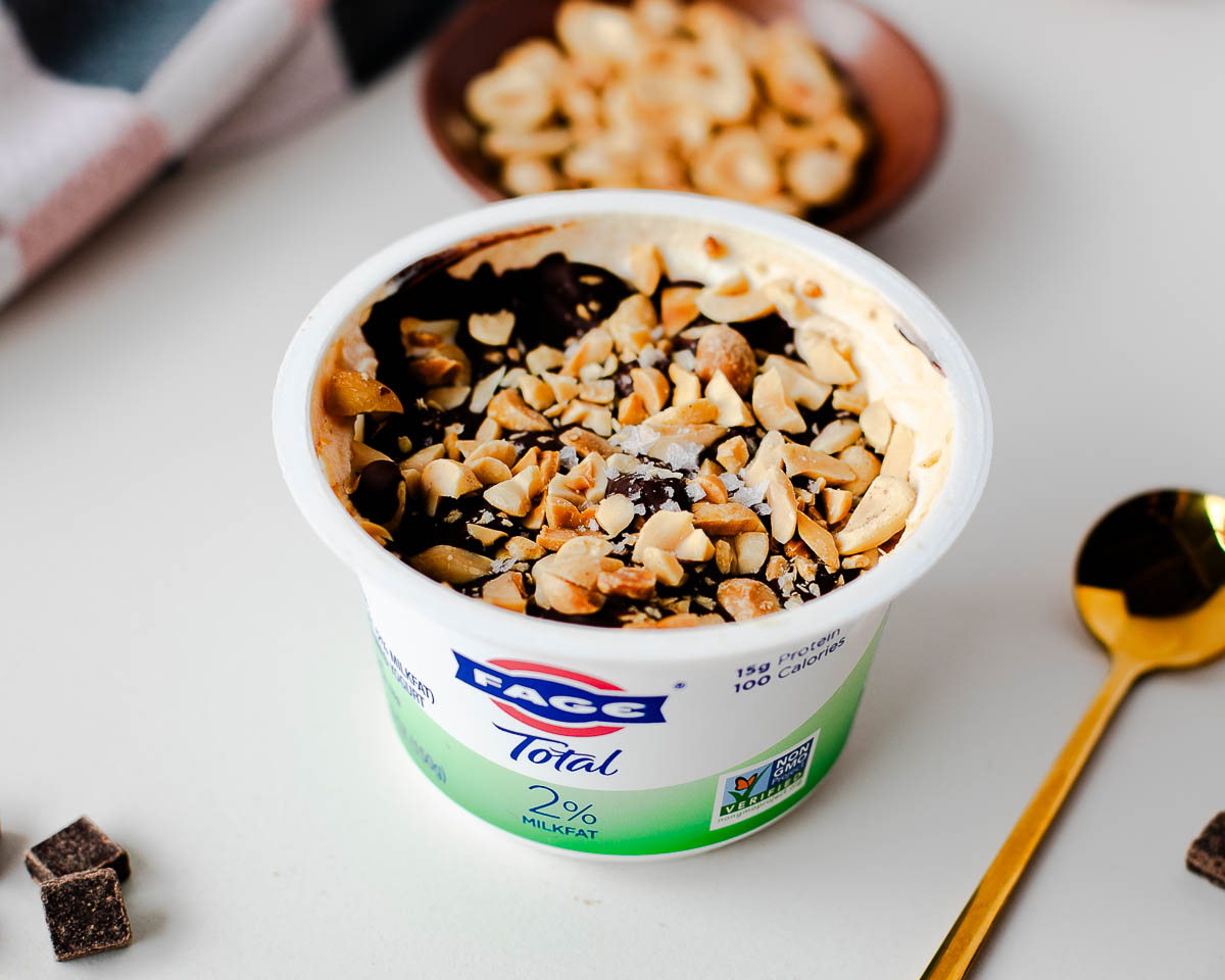 Snickers-inspired Greek yogurt cup with set chocolate shell and chopped peanuts, spoon alongside.