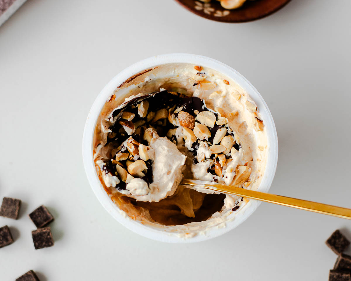 Spoon cracking the chocolate shell to reveal creamy peanut butter Greek yogurt and crunchy peanuts.