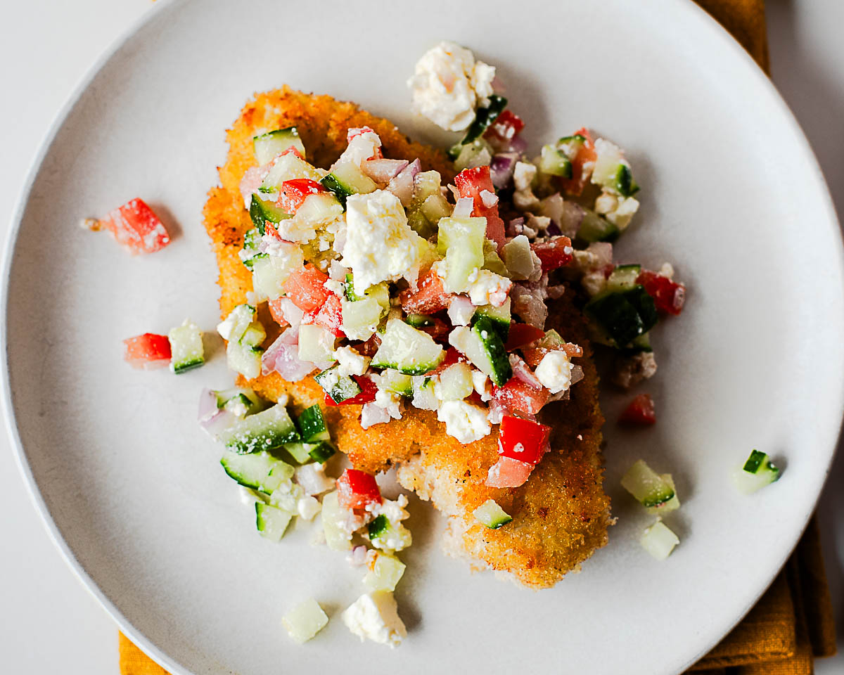 Overhead of crispy chicken cutlet piled with cucumber-tomato-feta salad.