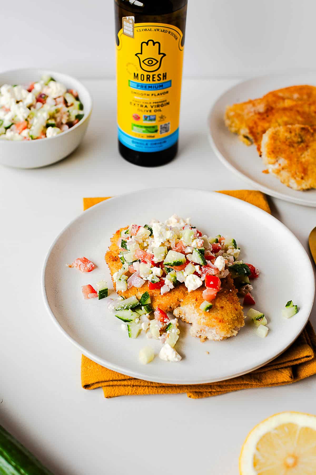 Crispy chicken cutlet topped with Mediterranean salad; Moresh olive oil bottle.
