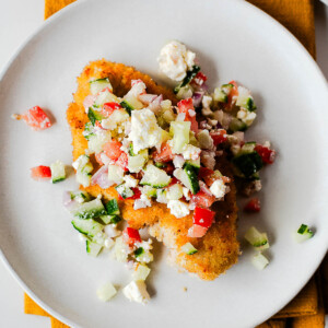 Close-up plate of crispy chicken cutlet with fresh Mediterranean topping.