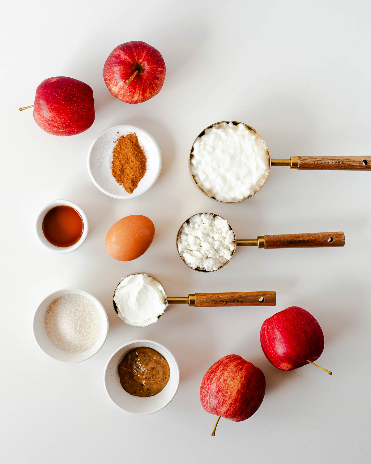 Ingredients for mini apple pie cheesecakes including fresh apples, cottage cheese, Greek yogurt, almond butter, vanilla, cinnamon, egg, and sugar measured out on a white surface.