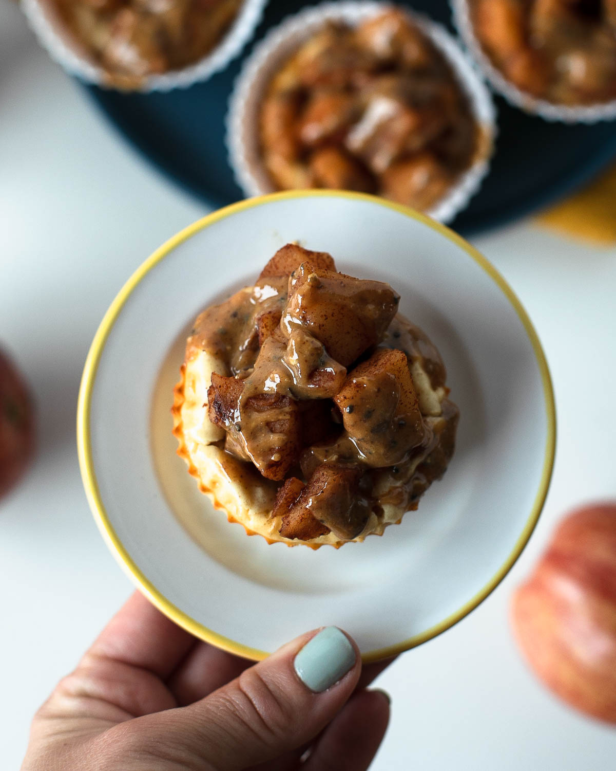 Hand holding a plate with a mini apple pie cheesecake topped with warm cinnamon apples and almond butter drizzle.