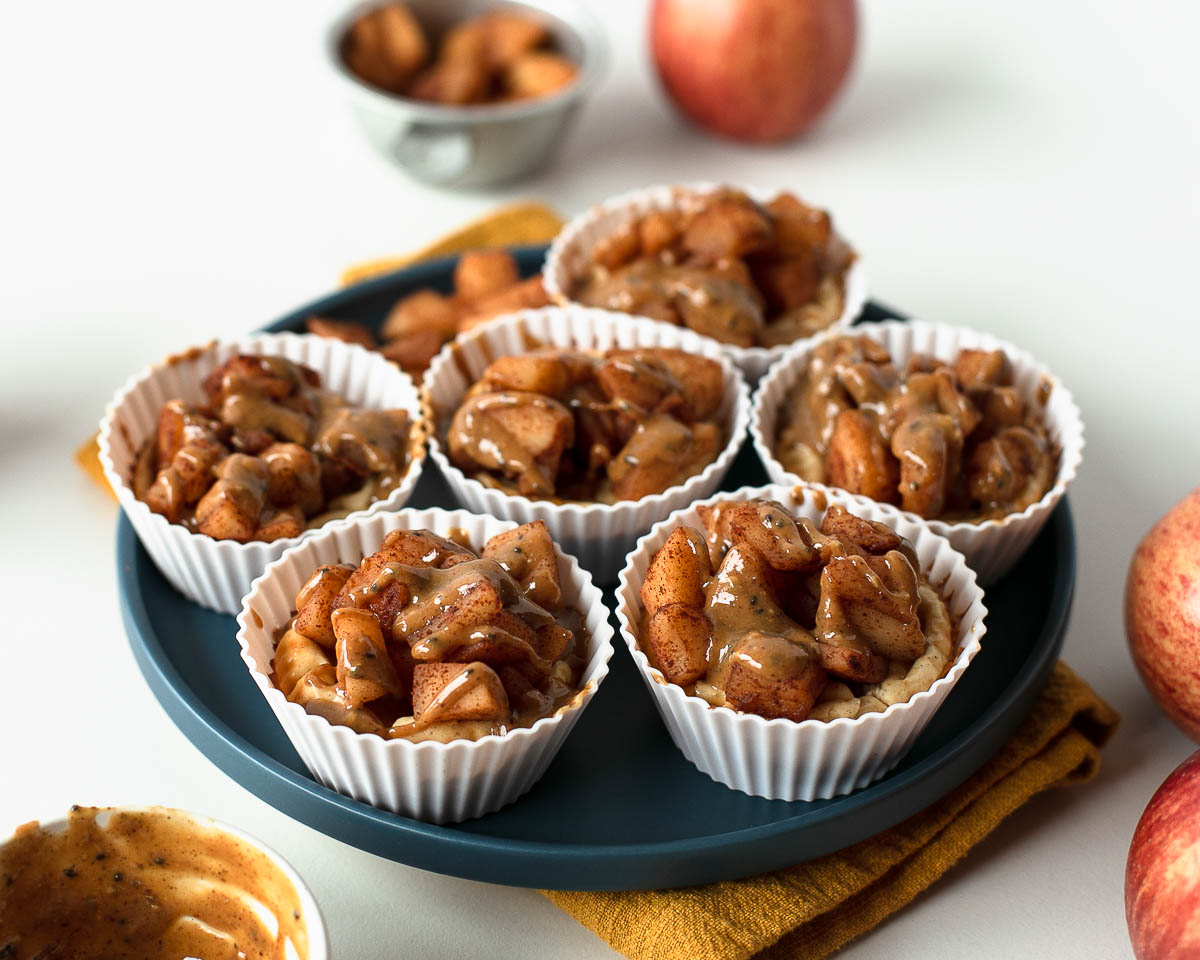 Six mini apple pie cheesecakes arranged on a blue plate, each topped with caramelized cinnamon apples and nutty maple drizzle.