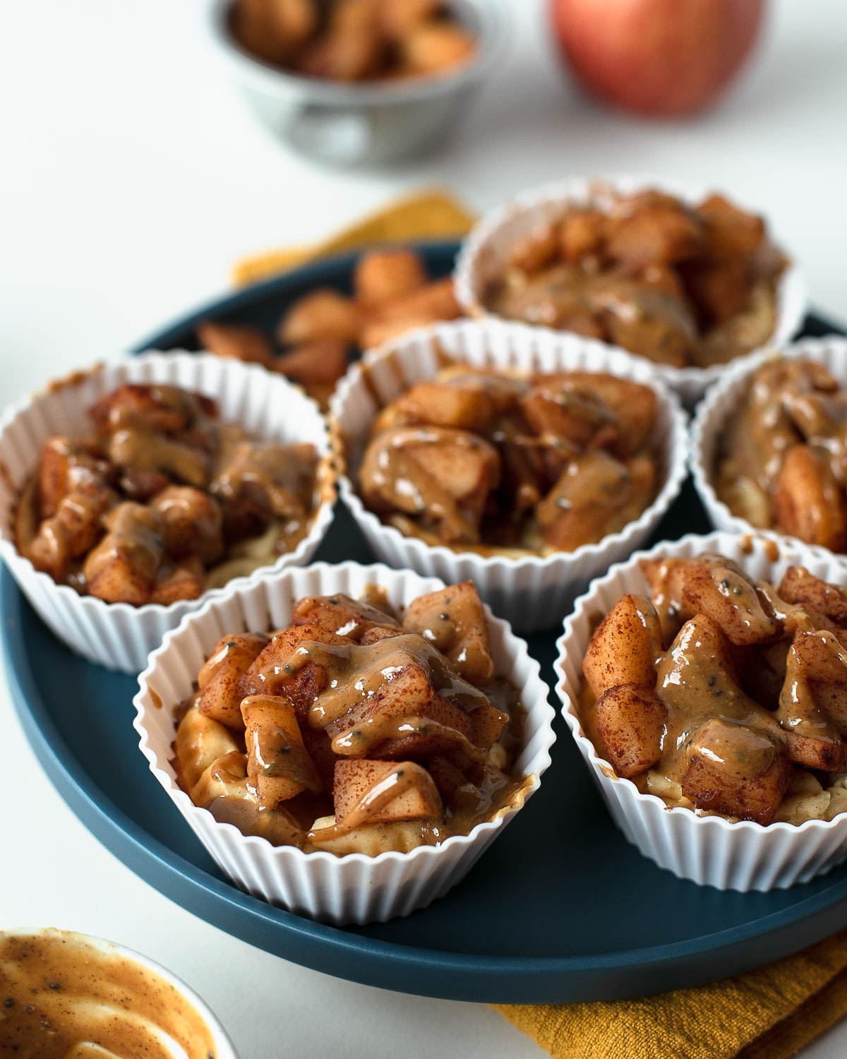 Close-up view of mini apple pie cheesecakes with golden apple topping and creamy drizzle, served on a plate with apples in the background.