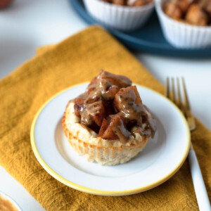 Single mini apple pie cheesecake on a small white plate with a golden rim, topped with spiced apples and drizzle, set on a mustard yellow napkin.