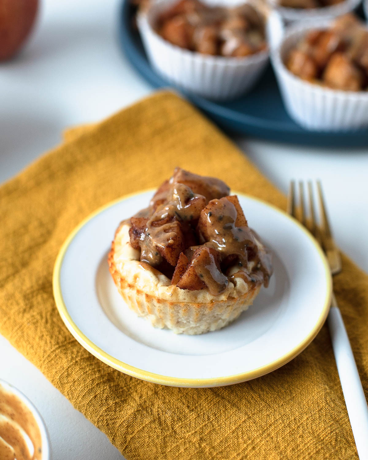 Single mini apple pie cheesecake on a small white plate with a golden rim, topped with spiced apples and drizzle, set on a mustard yellow napkin.