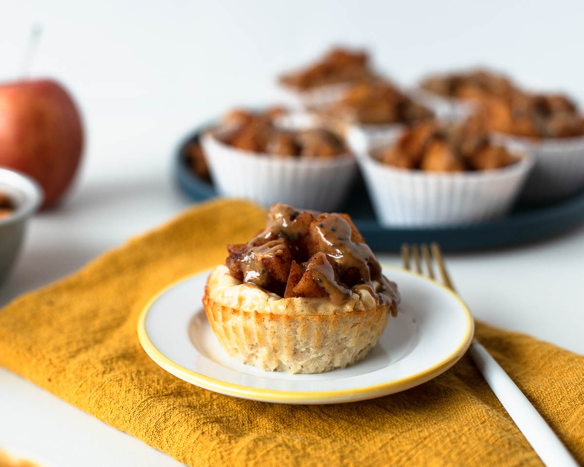 Mini apple pie cheesecake in focus on a plate, with more cheesecakes blurred in the background on a serving tray.