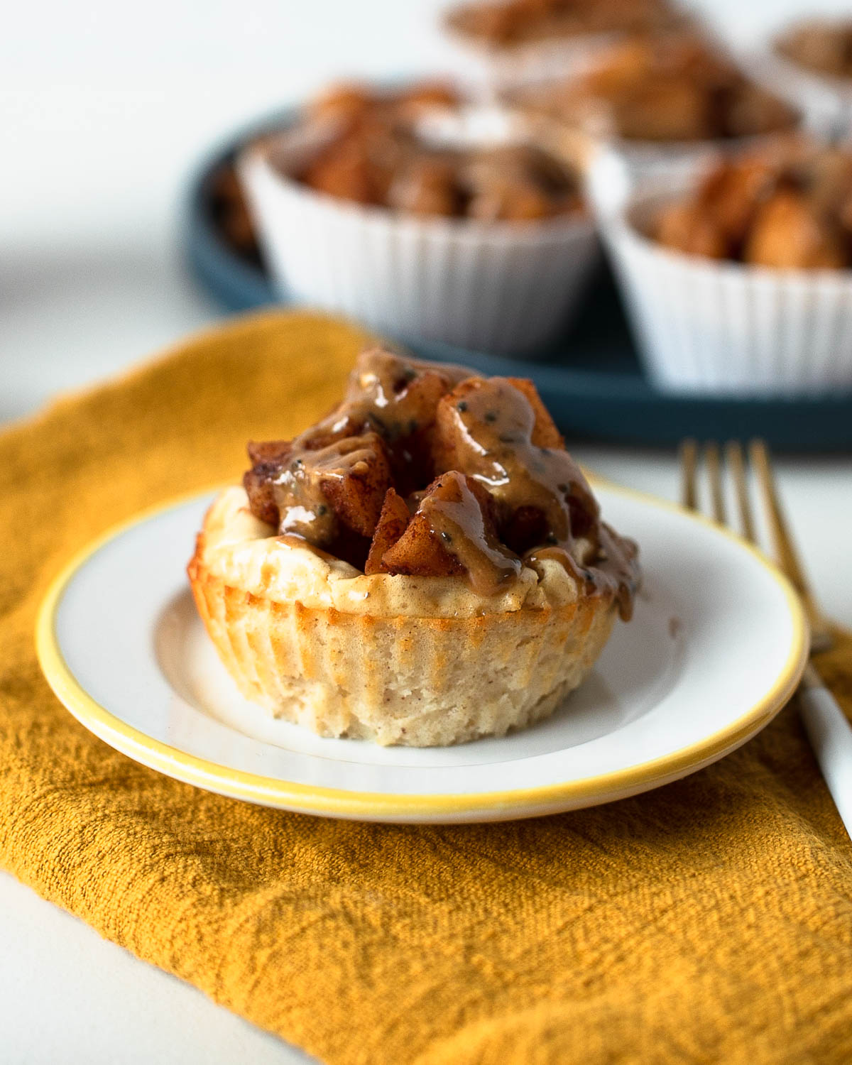 Close-up of a single mini apple pie cheesecake topped with warm cinnamon apples and almond butter drizzle on a yellow napkin with fork nearby.
