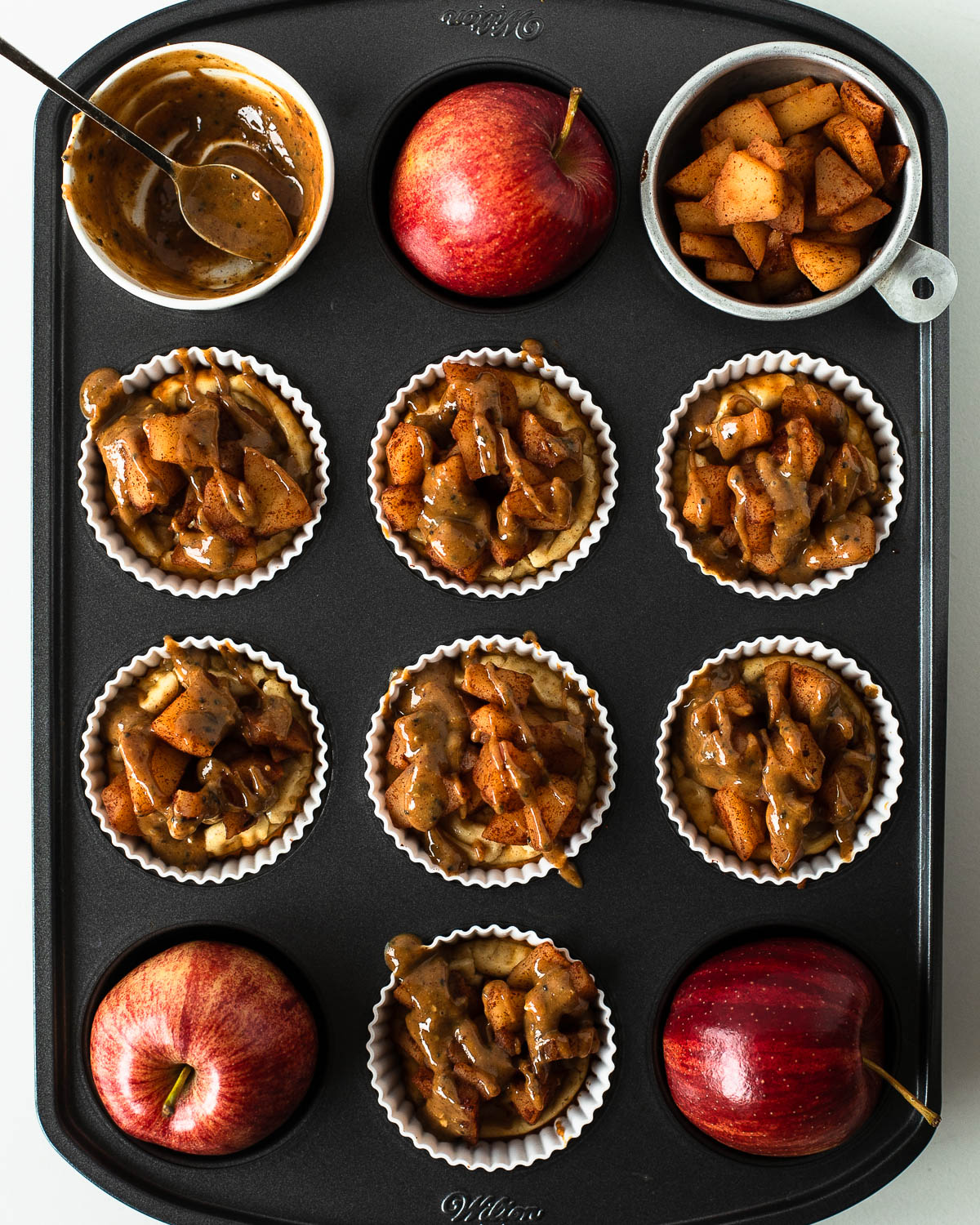 Mini apple pie cheesecakes topped with cinnamon apples and drizzled with almond butter maple sauce in a muffin tin with fresh apples on the side.