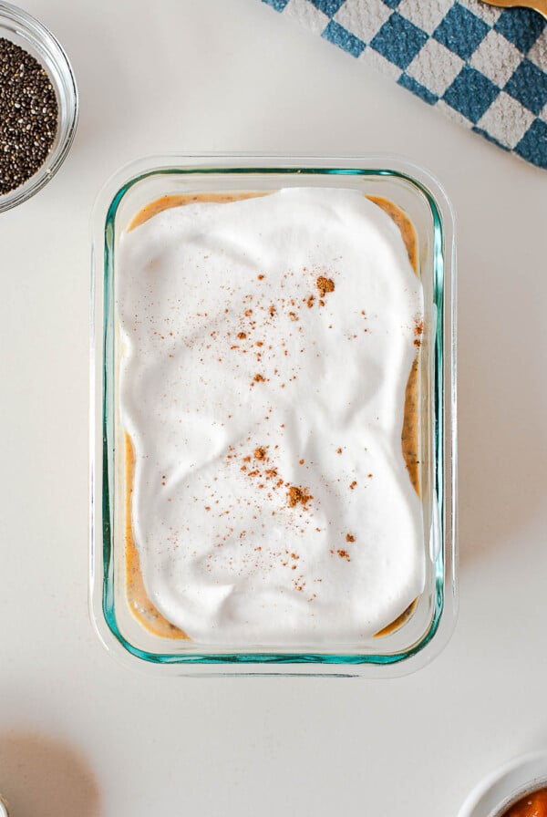 Pumpkin pie chia pudding topped with a layer of whipped coconut cream and a sprinkle of cinnamon in a glass dish, with chia seeds and a checkered towel nearby.
