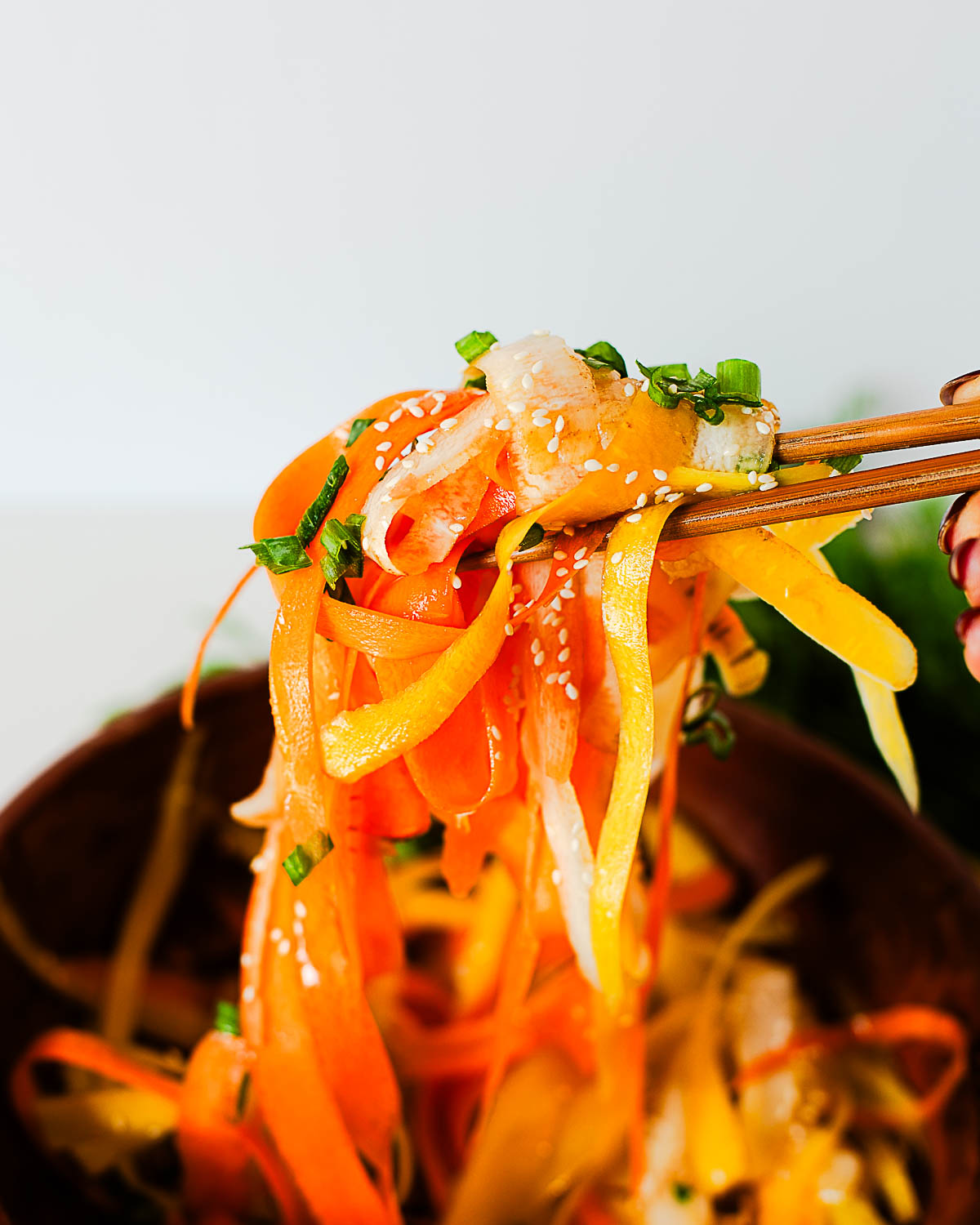 Chopsticks lifting carrot ribbons coated in olive oil dressing, showing sesame seeds and green onion garnish