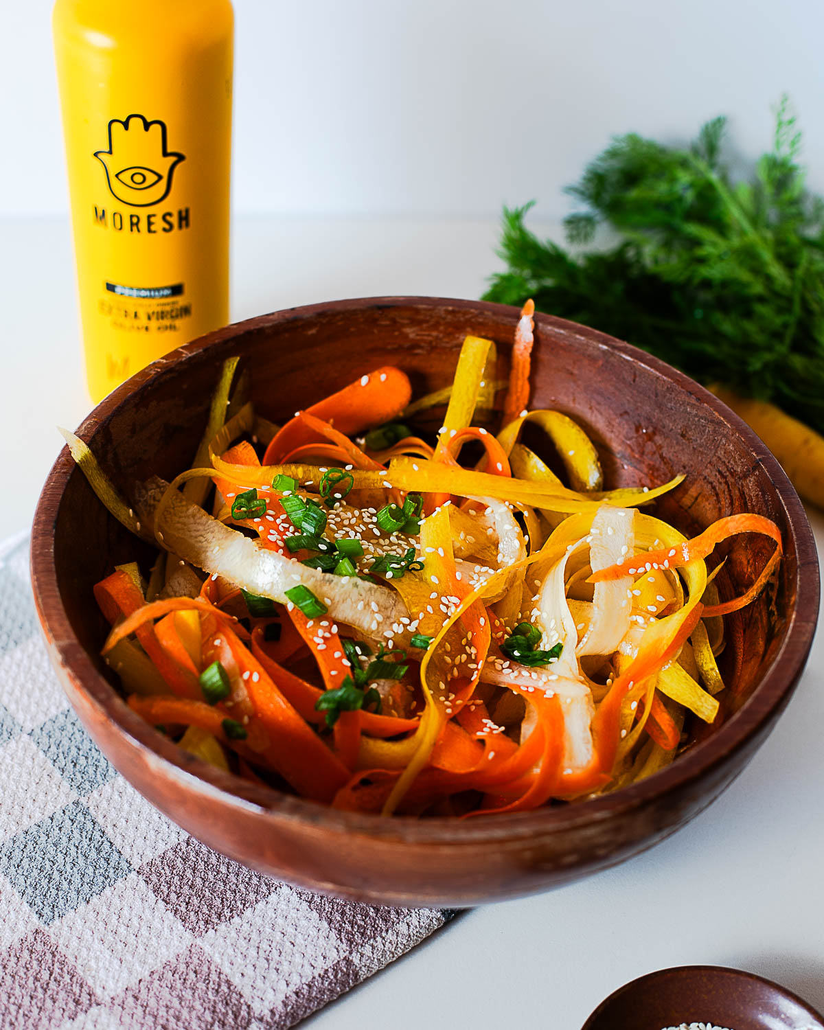 Carrot ribbon salad topped with green onions and sesame seeds, with a bottle of extra virgin olive oil in the background