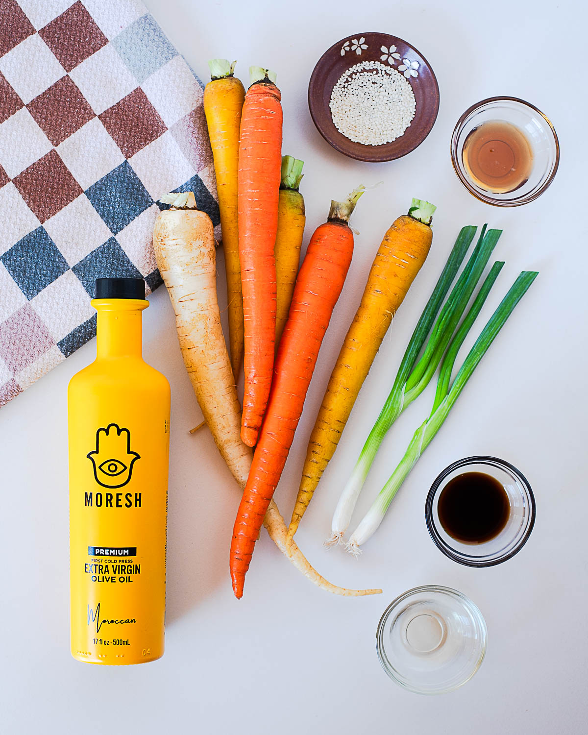 Carrot ribbon salad ingredients displayed: rainbow carrots, green onions, olive oil, rice vinegar, coconut aminos, maple syrup, and sesame seeds