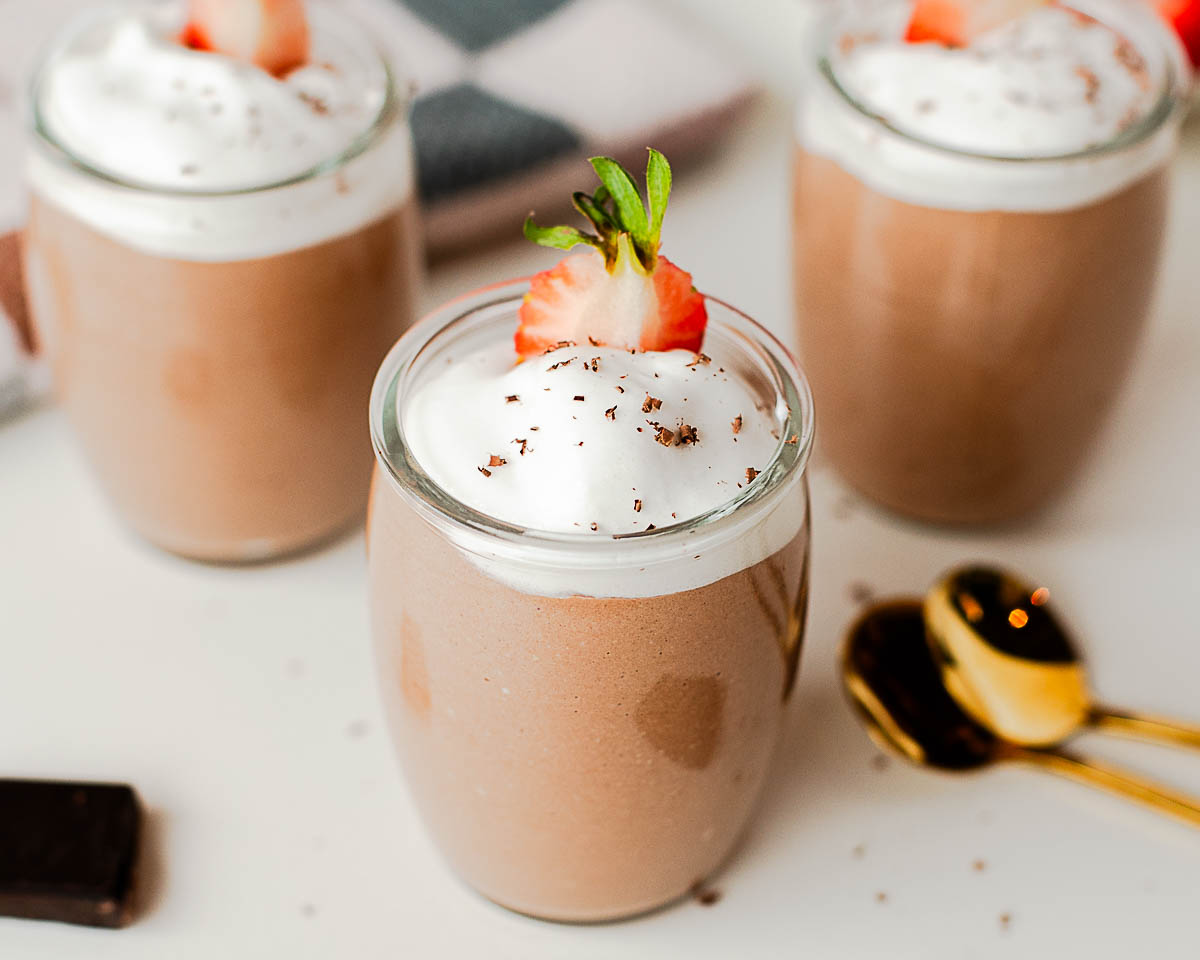 Multiple jars of cottage cheese chocolate pudding topped with whipped cream and strawberries on a light countertop.
