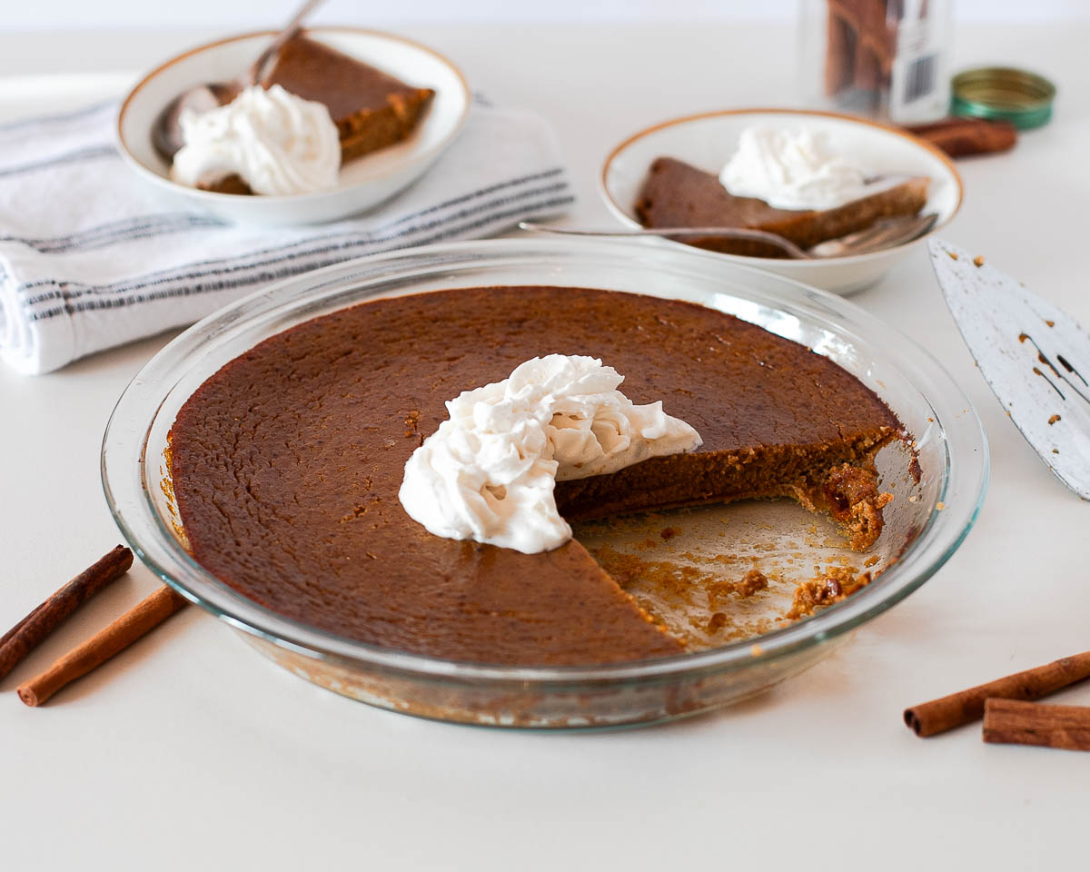 Crustless dairy free pumpkin pie in a glass dish with one slice removed, topped with whipped cream and surrounded by cinnamon sticks.
