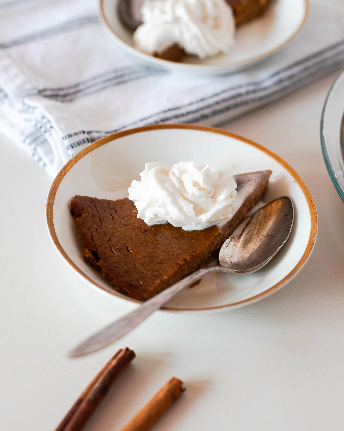 Close-up of a slice of crustless pumpkin pie on a white plate topped with whipped coconut cream, showing the creamy custard texture.