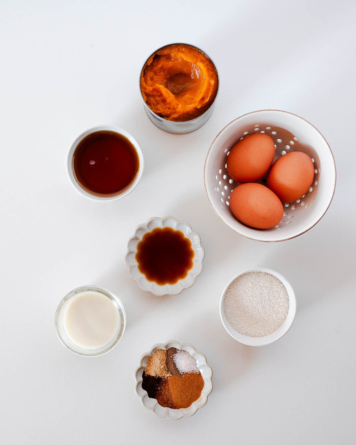Simple ingredients for a crustless dairy free pumpkin pie — pumpkin puree, eggs, maple syrup, non-dairy milk, sugar, vanilla, and warm fall spices arranged on a white surface.