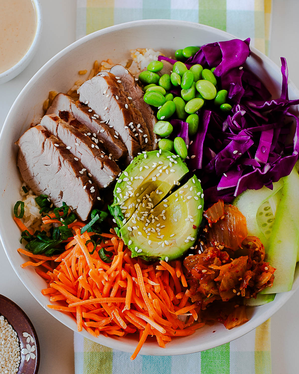 Colorful balanced bowl with sliced Asian marinated pork tenderloin, shredded carrots, purple cabbage, edamame, avocado, cucumber, kimchi, and sesame seeds over rice.