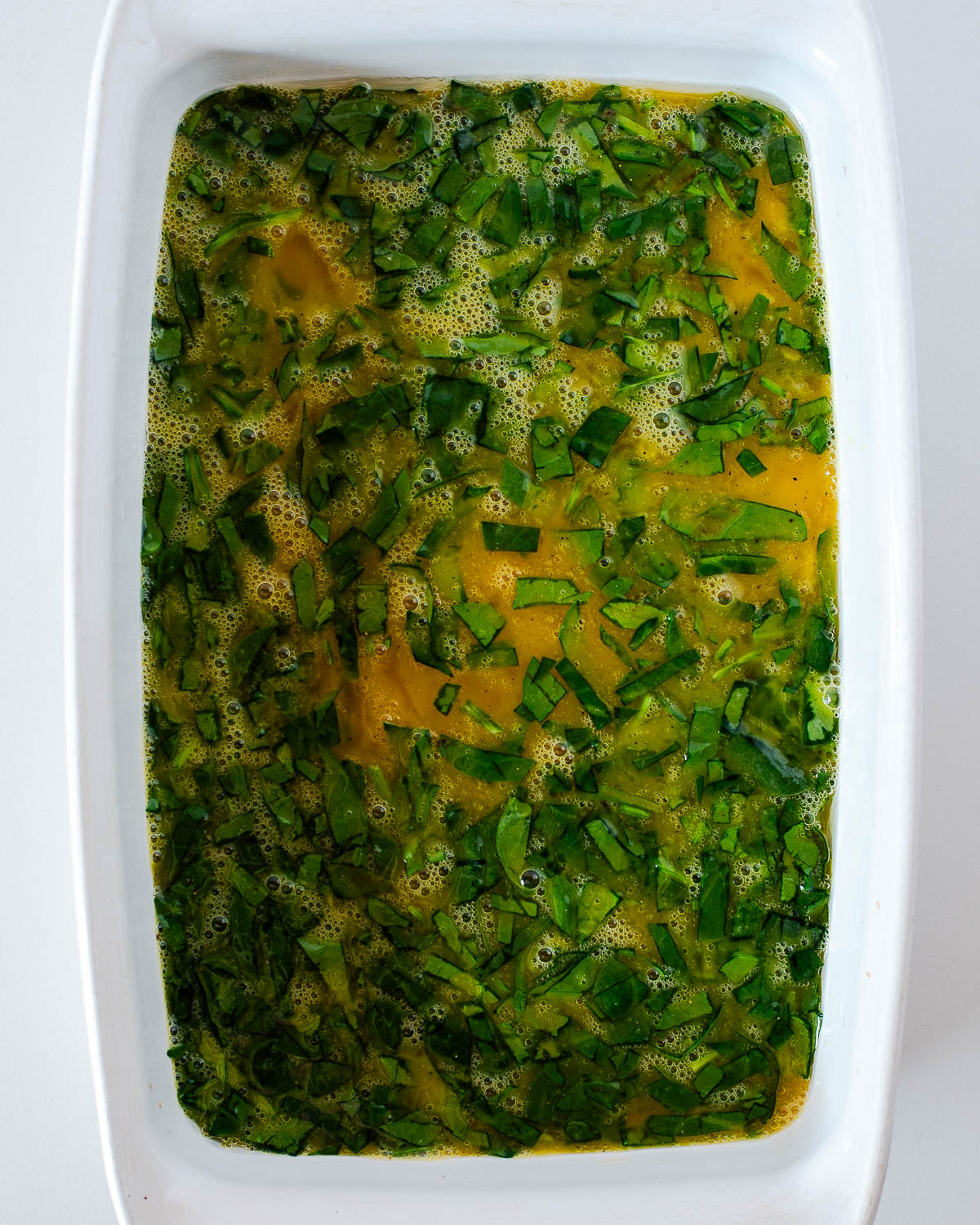 Whisked eggs and chopped spinach poured into a white baking dish, ready to bake for the breakfast sandwiches.