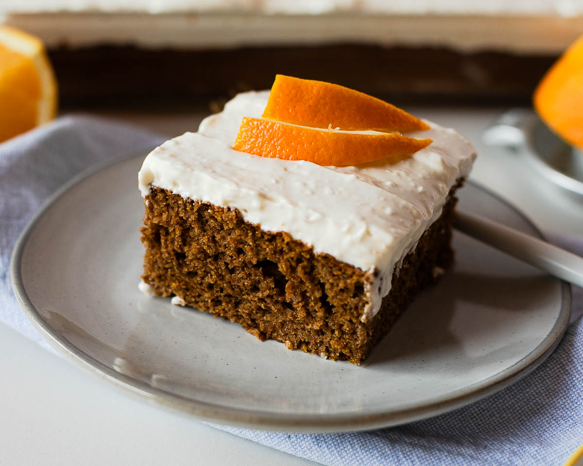 Close-up side view of a slice of sweet potato sheet cake topped with orange-scented cream cheese frosting and fresh orange peel garnish.