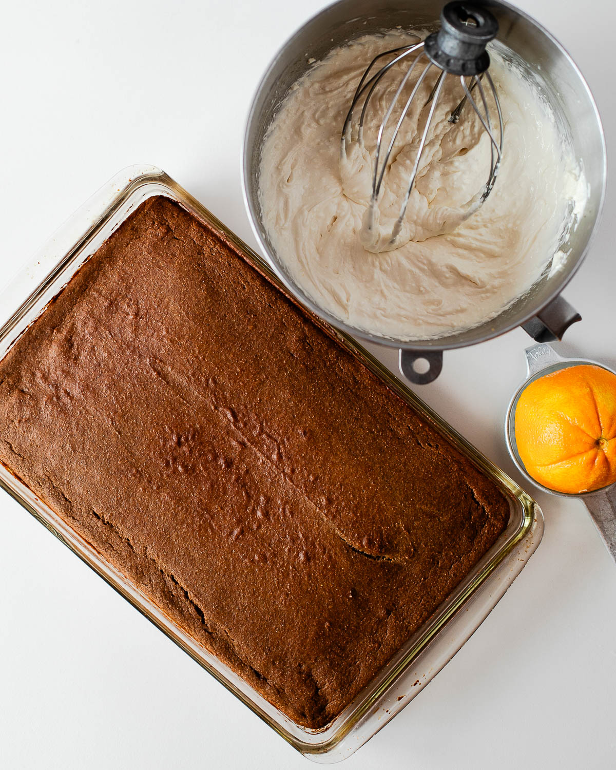 The cooled sweet potato cake next to a mixing bowl of orange-kissed cream cheese frosting and a fresh orange, ready to be frosted.