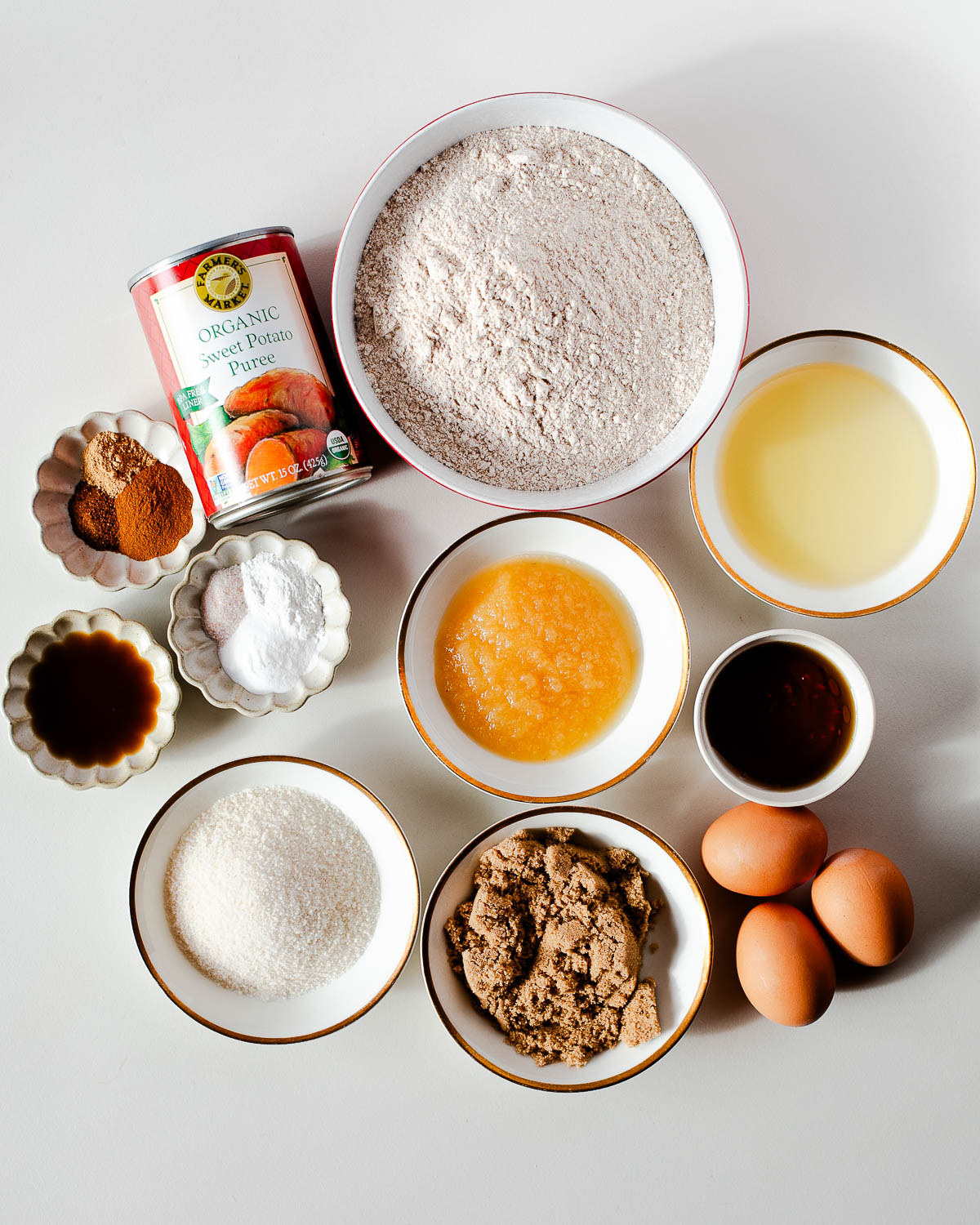 Overhead view of the cake ingredients arranged in small bowls — sweet potato puree, flour, sugars, eggs, applesauce, oil, vanilla, and warm spices.