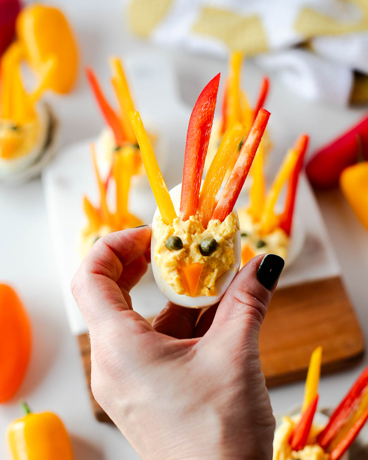 Hand holding a single turkey deviled egg decorated with red and yellow bell pepper feathers and caper eyes, with more turkey deviled eggs blurred in the background.