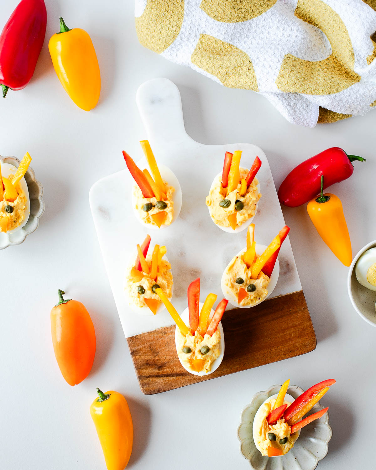 Colorful turkey deviled eggs on a white and wood serving board, decorated with red and yellow bell pepper “feathers” and caper eyes, surrounded by mini peppers.