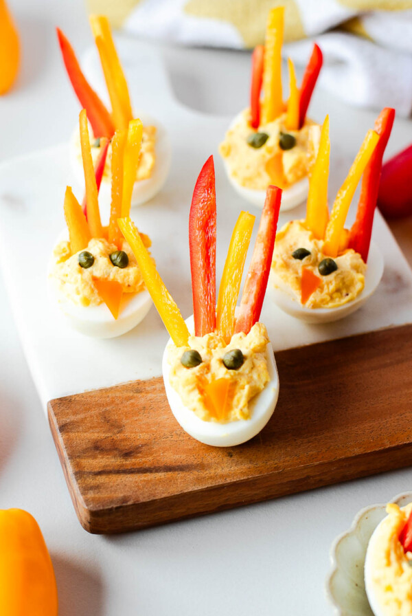 Front view of turkey deviled eggs on a white and wood board, each decorated with bright bell pepper feathers and a tiny orange pepper beak.