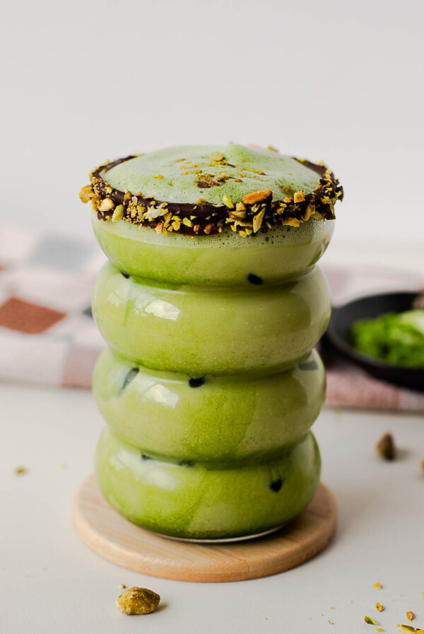 Iced pistachio protein matcha latte in a stacked glass with chocolate-pistachio rim on a wooden coaster.