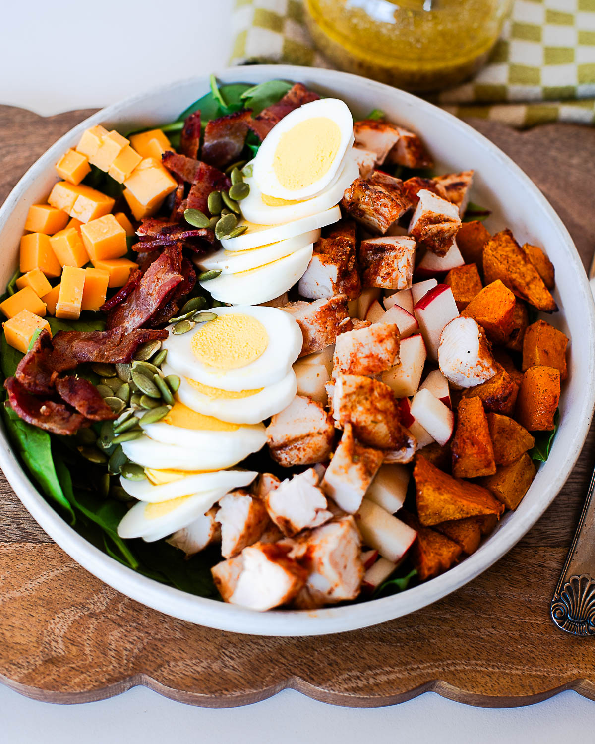 Harvest Cobb salad in a white bowl with spinach, sliced hard-boiled eggs, chopped Traeger chicken tenders, bacon, cheddar cubes, apples, roasted sweet potatoes, pumpkin seeds, and dressing.