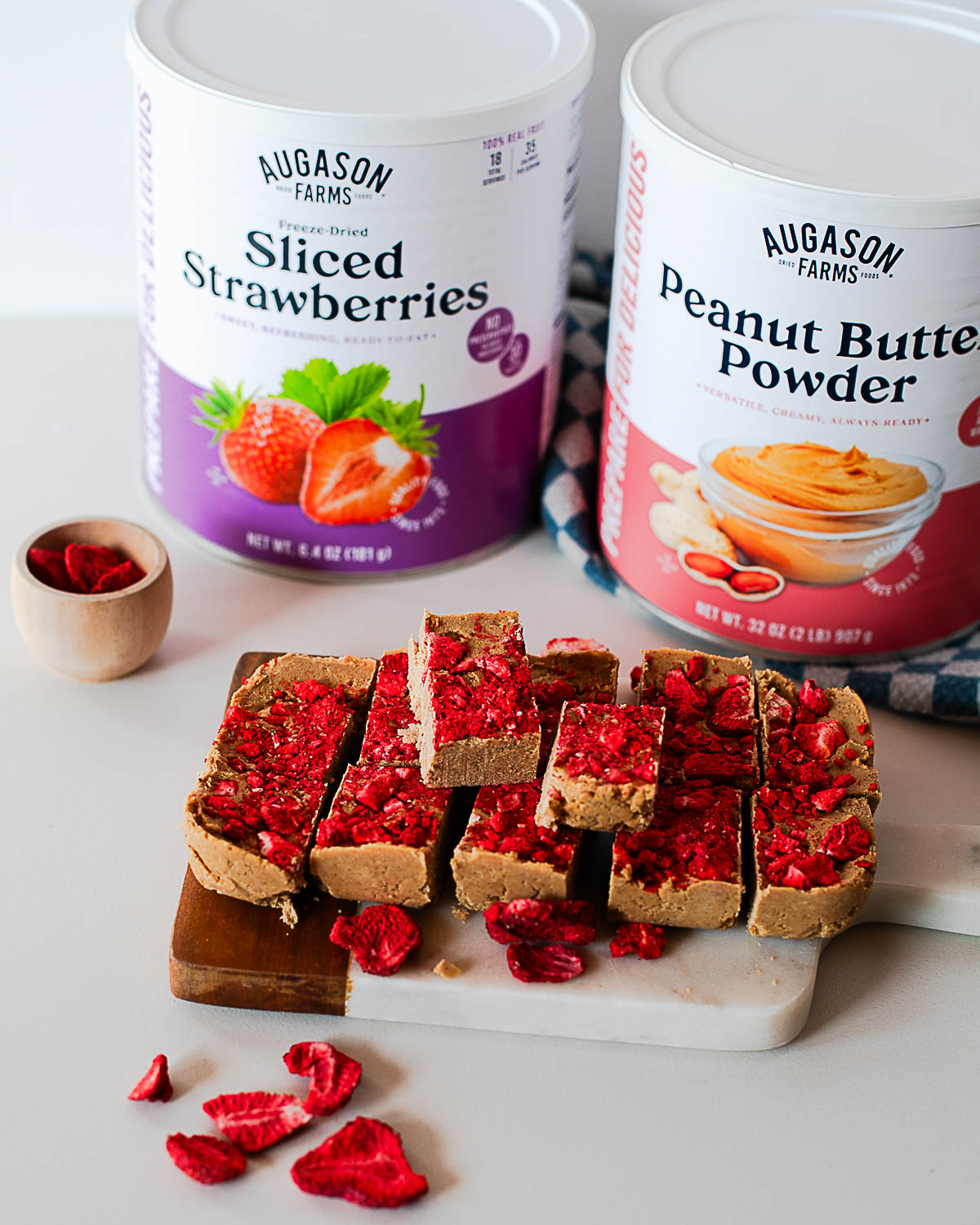 Sliced PB&J protein fudge squares topped with freeze-dried strawberries, with Augason Farms peanut butter powder and freeze-dried strawberries in the background.