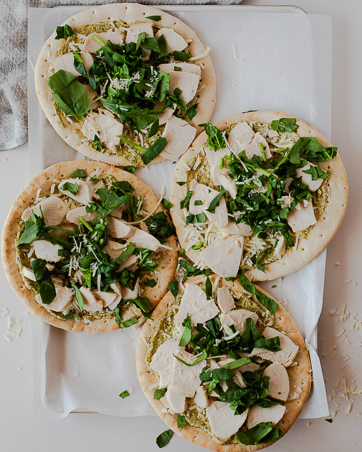 Overhead view of four pita flatbread pizzas topped with creamy pesto sauce, shredded mozzarella, sliced chicken, and fresh spinach on a parchment-lined baking sheet.